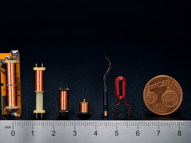EMG_Sortiment_wickelei Vor einem schwarzen Hintergrund sind eine Reihe kleiner Kupferspulen und elektronische Bauteile unterschiedlicher Größe neben einer 5-Eurocent-Münze und einem Lineal als Maßstab angeordnet.