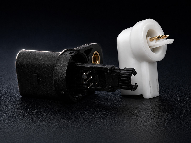 Detailaufnahme von elektrischen Steckverbindern Zwei elektronische Bauteile – eines schwarz und eines weiß – mit Metallanschlüssen liegen auf einer dunklen, strukturierten Oberfläche. Das schwarze Bauteil liegt flach, während das weiße aufrecht steht; bei beiden sind die Anschlussenden zu sehen.